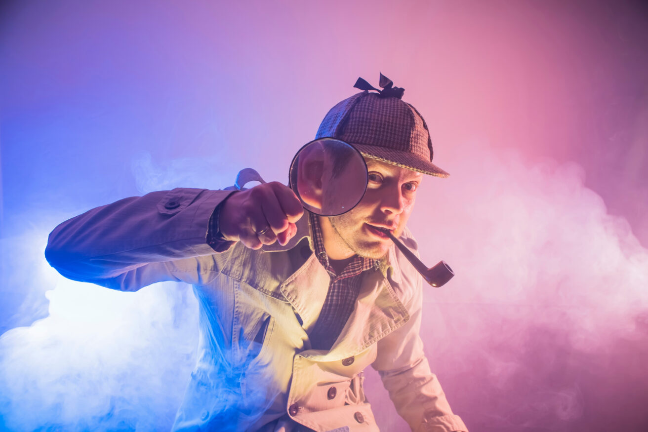 Man dressed as detective peering into camera with magnifying glass in front of pink and blue smokey backdrop.